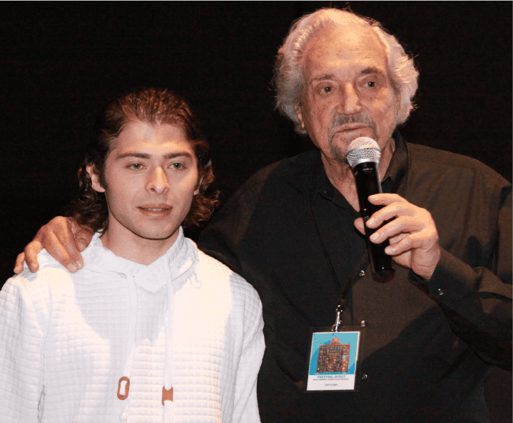 Elderly man and young man speaking into a microphone, introducing a film for the Toronto Jewish Film Festival